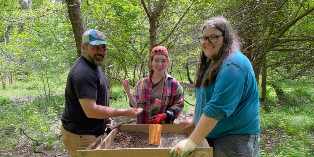 Three people smile while sifting soil at an outdoor archaeology dig in a wooded area in collaboration with ARI