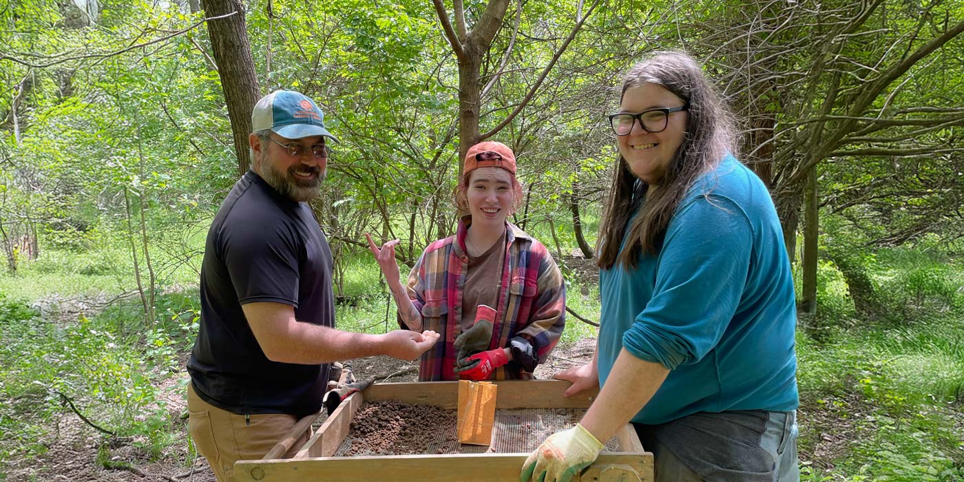 Three people smile while sifting soil at an outdoor archaeology dig in a wooded area in collaboration with ARI