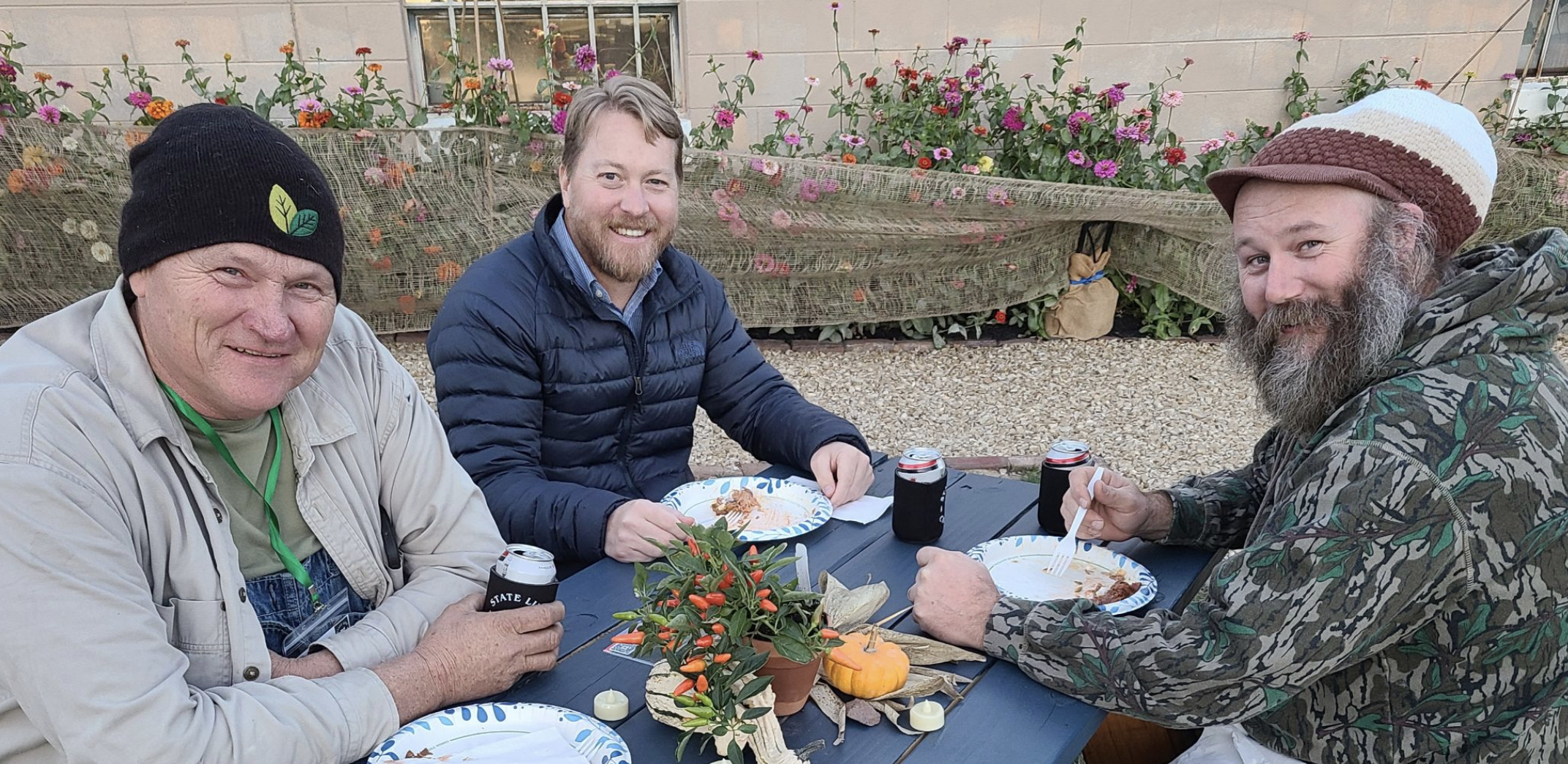 Three smiling men sit at a picnic table outdoors, enjoying plates of food with fall-themed table decorations and flowers in the background.