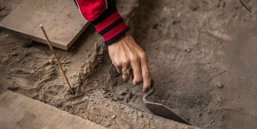 Close-up of a person carefully digging in soil with a small archaeological trowel at an excavation site