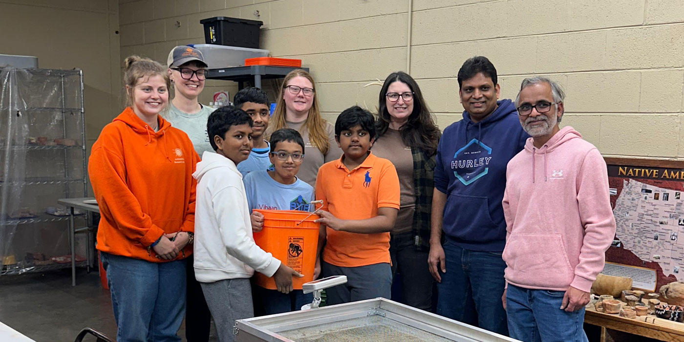 FIRST LEGO League students and ARI staff pose together during a hands-on archaeology learning experience, showcasing a student-designed excavation tool as part of a STEM education partnership