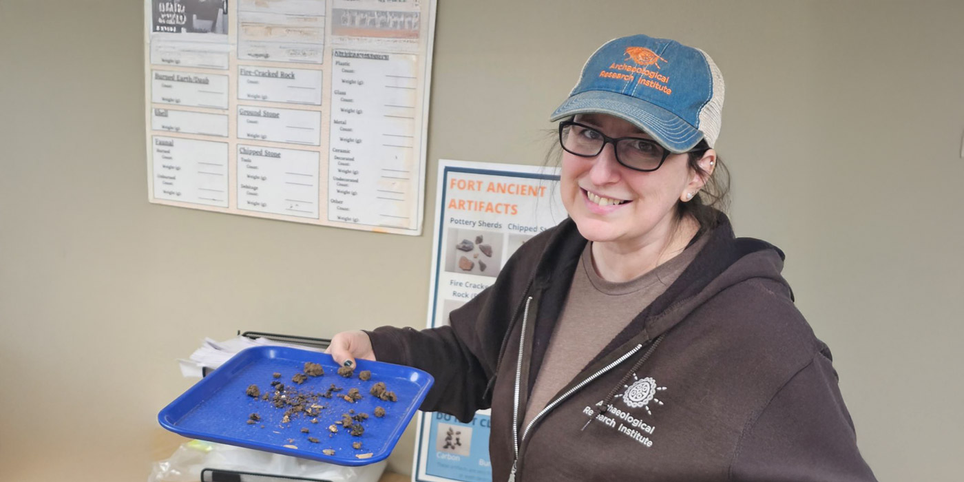 Archaeologist holding a blue tray of soil and artifact fragments during winter lab processing at an archaeology research lab.